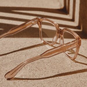 Close-up of QUAY’s transparent blush pink square eyeglasses with thick frames resting on a textured concrete surface in warm sunlight.
