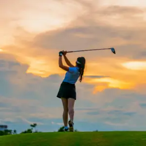 Woman golfer in a blue shirt and black skirt swings a golf club on a grassy hill at sunset.