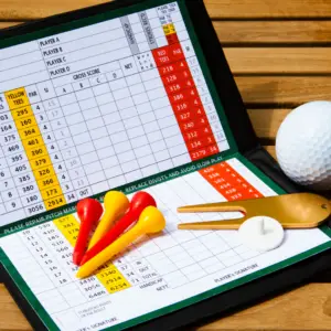 Close-up of a golf scorecard on a clipboard with yellow, red, and orange tees, a white golf ball, and a brass divot tool on a wooden table.
