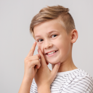 Smiling boy inserts a contact lens into his right eye while looking at the camera in a striped shirt.
