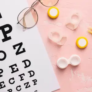 Eye chart on a white sheet with black letters, alongside pink textured background and contact-lens accessories (glasses, lens cases) scattered nearby.