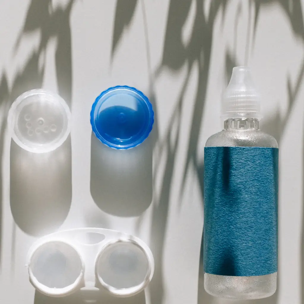Open contact lens case with two lenses and lids nearby, plus a bottle of blue-labeled contact lens solution on a white surface.